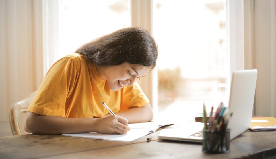 Student writing notes at a desk with a laptop
